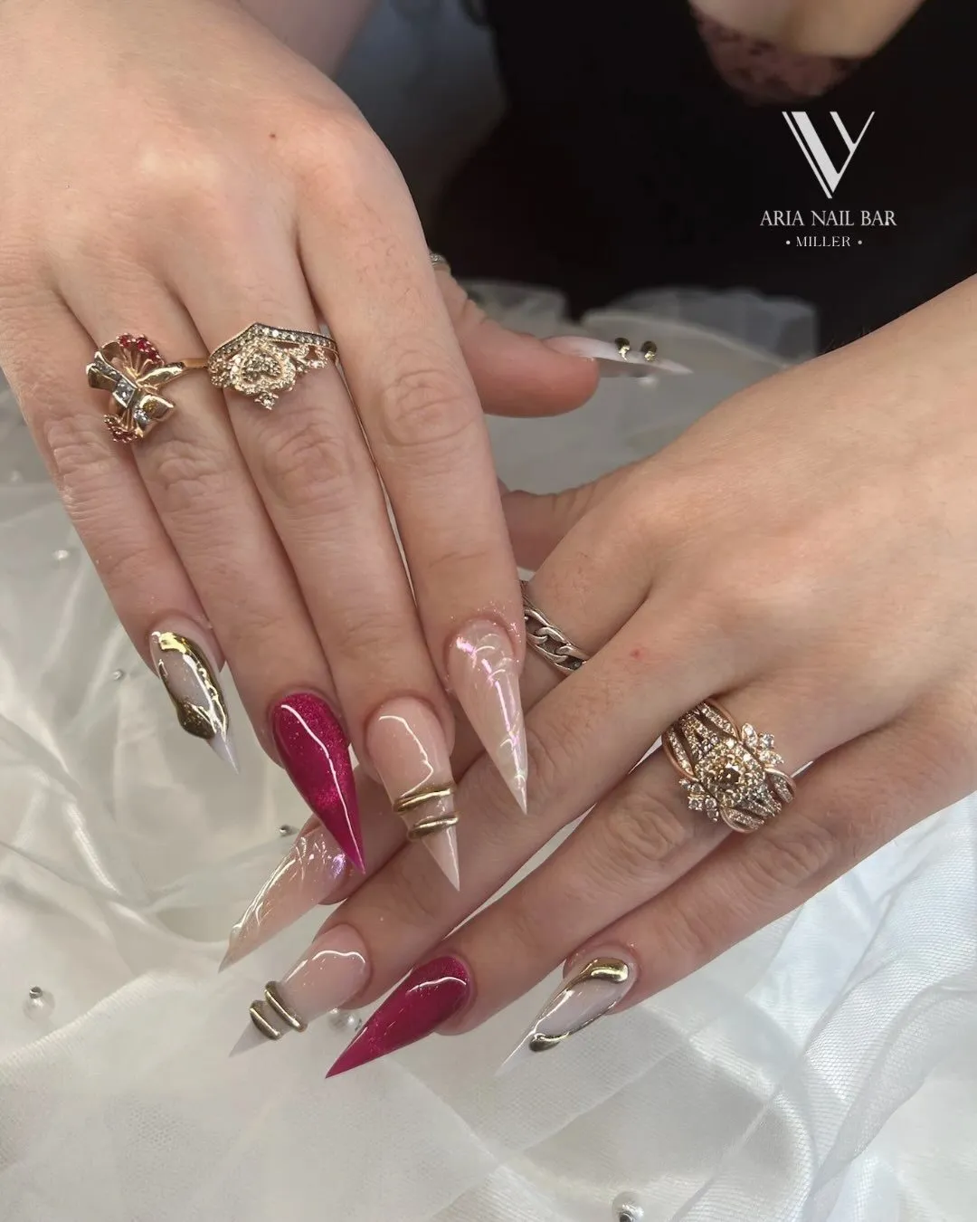 Long stiletto nails in clear, burgundy and gold foil, styled with stacked gold rings.